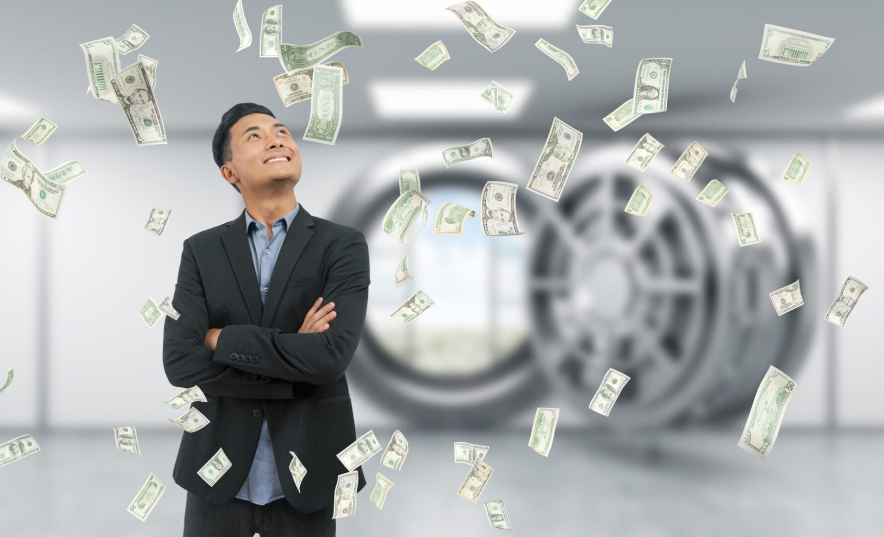 Businessman in suit standing in bank vault with hands folded, smiling. Door open. City view seen through it. Dollar notes are falling at him. Concept of successful business idea implementation.